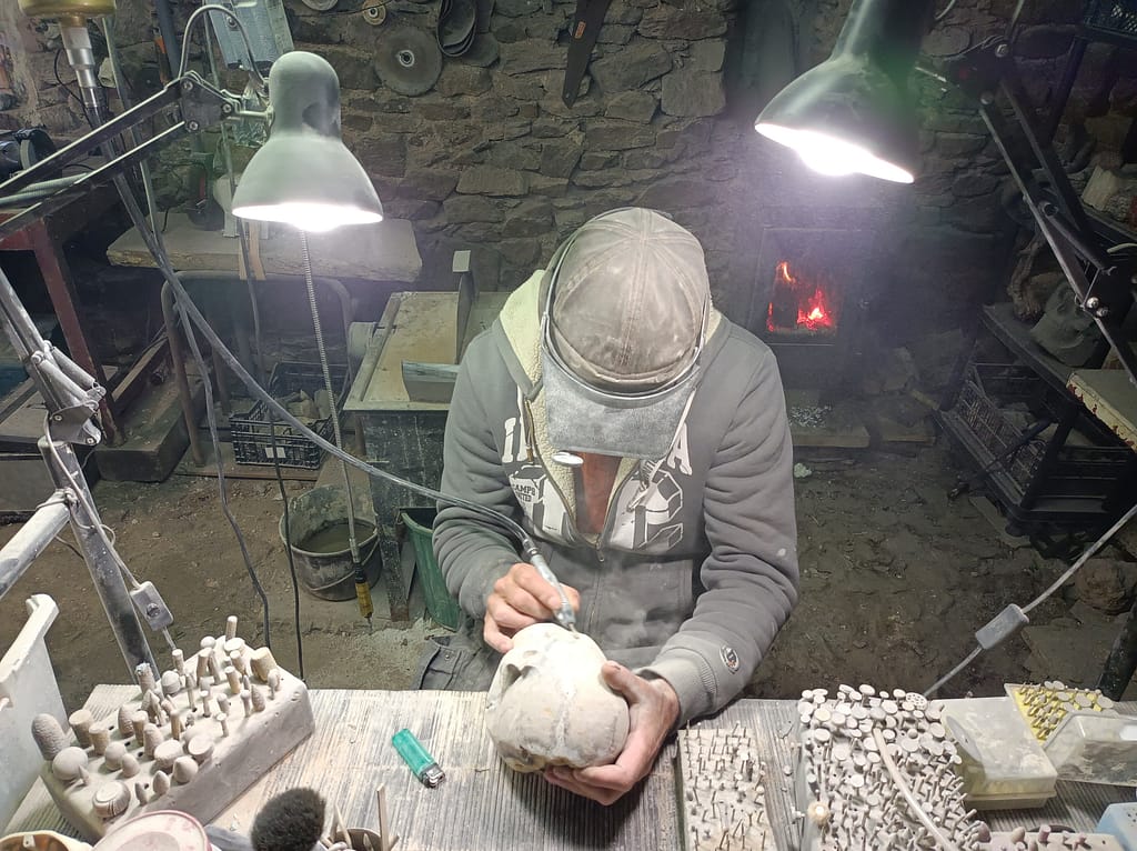 Atelier Karving12 : sculpteur en travail, façonnant à la main un crâne en septaria, travail artisanal sur établi avec outils de taille, scène authentique de création en atelier.