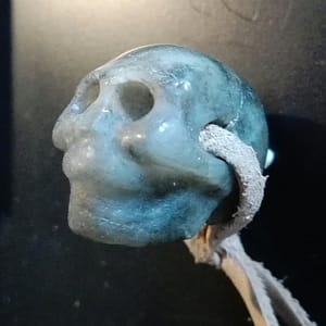 Pendentif crâne sculpté en labradorite, vue de profil, sculpture minérale artisanale en pierre naturelle aux reflets bleus et verts, pièce unique montée en pendentif, création d’art minéral présentée en galerie.