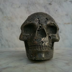 Crâne sculpté en pyrite, vue de face, sculpture minérale en pierre naturelle, cristallisations de pyrite visibles, orbites creusées et mâchoire détaillée, pièce d’art minéral exposée en galerie.
