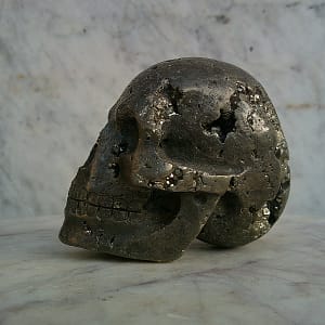 Crâne sculpté en pyrite, vue de profil, sculpture minérale en pierre naturelle cristallisée, détails des cavités et cristaux visibles, pièce d’art présentée en galerie.