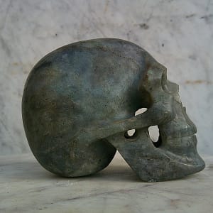 Vue de profil gauche d’un crâne sculpté à la main en labradorite polie, mettant en évidence les volumes du crâne et la structure minérale naturelle. Sculpture minérale artisanale en labradorite.