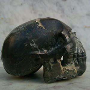 Vue de profil gauche d’un petit crâne sculpté en pierre magnétique entièrement polie, révélant la ligne du crâne, l’arcade sourcilière et la mâchoire dans un seul bloc minéral. Sculpture taillée à la main.
