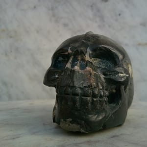Vue frontale d’un petit crâne sculpté en pierre magnétique entièrement polie, mettant en évidence le travail précis des orbites, de la mâchoire et de la dentition. Sculpture minérale taillée à la main.