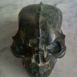Petit crâne sculpté à la main dans une pierre magnétique d’origine météoritique, vue frontale mettant en évidence la crête centrale sculptée sur le sommet du crâne, sculpture minérale format miniature, taillée et polie en atelier, avec ligne de crête volontairement marquée, reliefs anguleux, orbites creusées et textures métalliques naturelles de la pierre, pièce unique.