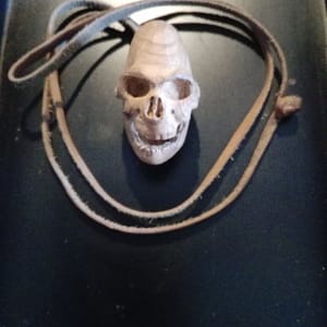 Crâne miniature sculpté à la main dans une loupe de bois, vue de face, pendentif artisanal monté sur lien en cuir, mettant en valeur les volumes du visage, les orbites creusées, la mâchoire détaillée et le grain naturel du bois, sculpture organique issue d’un travail de taille fine en atelier, pièce unique présentée en galerie, non proposée à la vente.