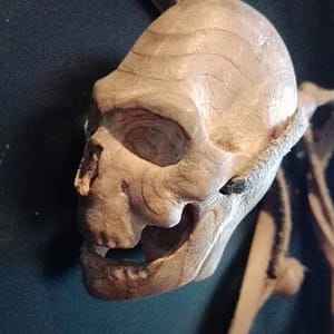 Petit crâne sculpté à la main dans une loupe de bois, pendentif artisanal présenté en galerie, sculpture miniature mettant en valeur la texture naturelle du bois, les veines organiques et le travail de taille fine, bijou sculptural unique, photographié porté en pendentif, pièce artistique non destinée à la vente.