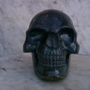Sculpture de crâne humain en pyrite, vue frontale centrée, pièce unique sculptée à la main dans un bloc de minéral naturel, mettant en évidence la symétrie du visage, la profondeur des orbites, la dentition détaillée et les reflets métalliques caractéristiques de la pyrite travaillée à l’atelier.