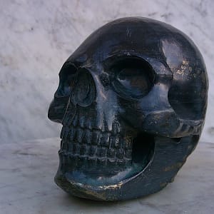 Sculpture de crâne humain en pyrite, vue de profil gauche, pièce unique taillée à la main dans un bloc de minéral naturel, mettant en évidence la structure cristalline de la pyrite, les reflets métalliques et le travail précis des volumes réalisé à l’atelier.