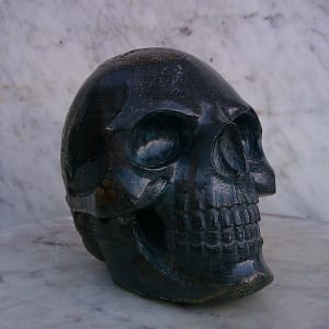 Sculpture de crâne humain en pyrite, vue de trois-quarts face, pièce unique sculptée à la main dans un bloc de minéral naturel, mettant en évidence les orbites, la dentition finement travaillée, les reflets métalliques et les variations de couleur propres à la pyrite façonnée à l’atelier.