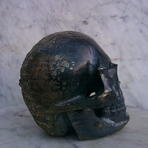 Sculpture de crâne humain en pyrite, vue de trois-quarts arrière opposé, pièce unique sculptée à la main dans un bloc de minéral naturel, mettant en évidence les volumes arrière du crâne, les zones de pyrite laissées plus brutes, les inclusions métalliques et les traces du geste de sculpture réalisées à l’atelier.