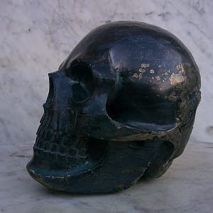 Sculpture de crâne humain en pyrite, vue de profil gauche opposé, pièce unique sculptée à la main dans un bloc de minéral naturel, mettant en évidence les variations de texture de la pyrite et les reflets métalliques liés à la structure minérale travaillée à l’atelier.