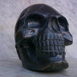 Sculpture de crâne humain en pyrite, vue de face légèrement de trois-quarts, pièce unique sculptée à la main dans un bloc de minéral naturel, révélant les reflets métalliques, les inclusions de pyrite et le travail fin des détails anatomiques réalisé à l’atelier.