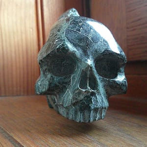 Sculpture de crâne en serpentine, vue de face légèrement de trois-quarts, pièce unique sculptée à la main dans un bloc de minéral naturel, présentée en galerie comme exemple de travail de sculpture minérale, mettant en évidence les orbites profondes, la structure veineuse de la serpentine, les zones polies et les traces du façonnage des volumes à l’atelier.