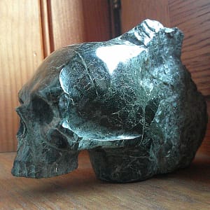 Sculpture de crâne en serpentine, vue de profil, pièce unique sculptée à la main dans un bloc de minéral naturel, présentée en galerie comme exemple de travail de sculpture minérale, mettant en évidence la texture de la serpentine, les veines internes et le façonnage des volumes à l’atelier.